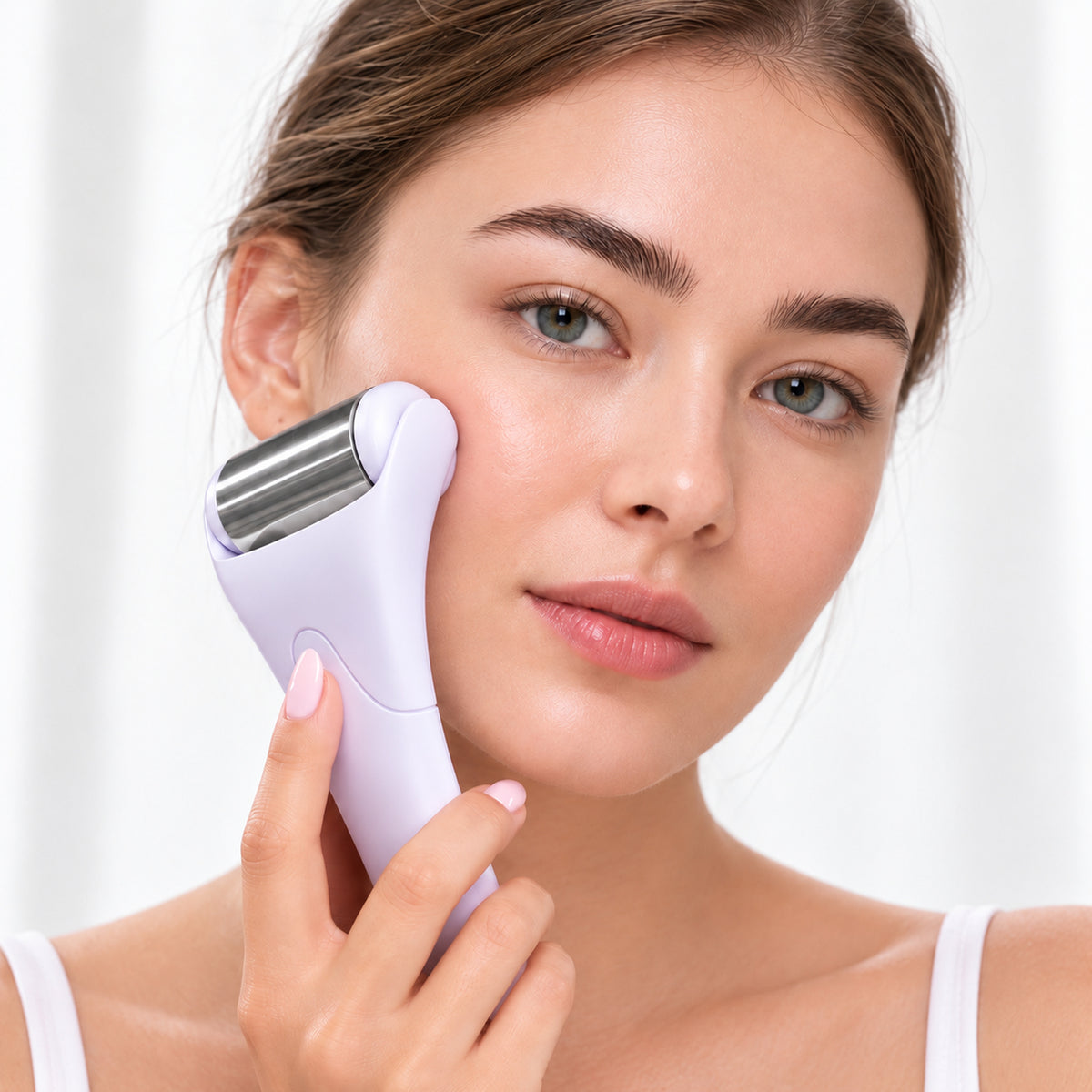 A close-up of a woman using a light purple handheld facial skin device with a silver metal roller on her cheek, set against a bright, soft-focus background.